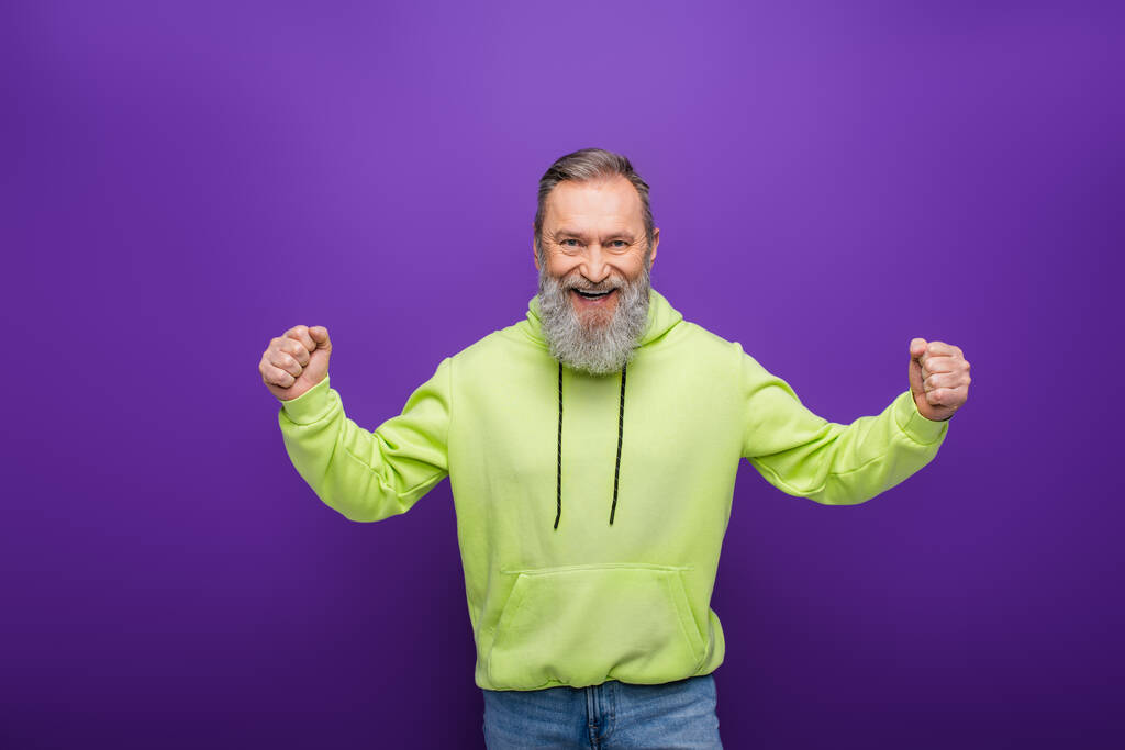 excited bearded man with grey hair wearing green hoodie and gesturing on purple  - Photo, Image