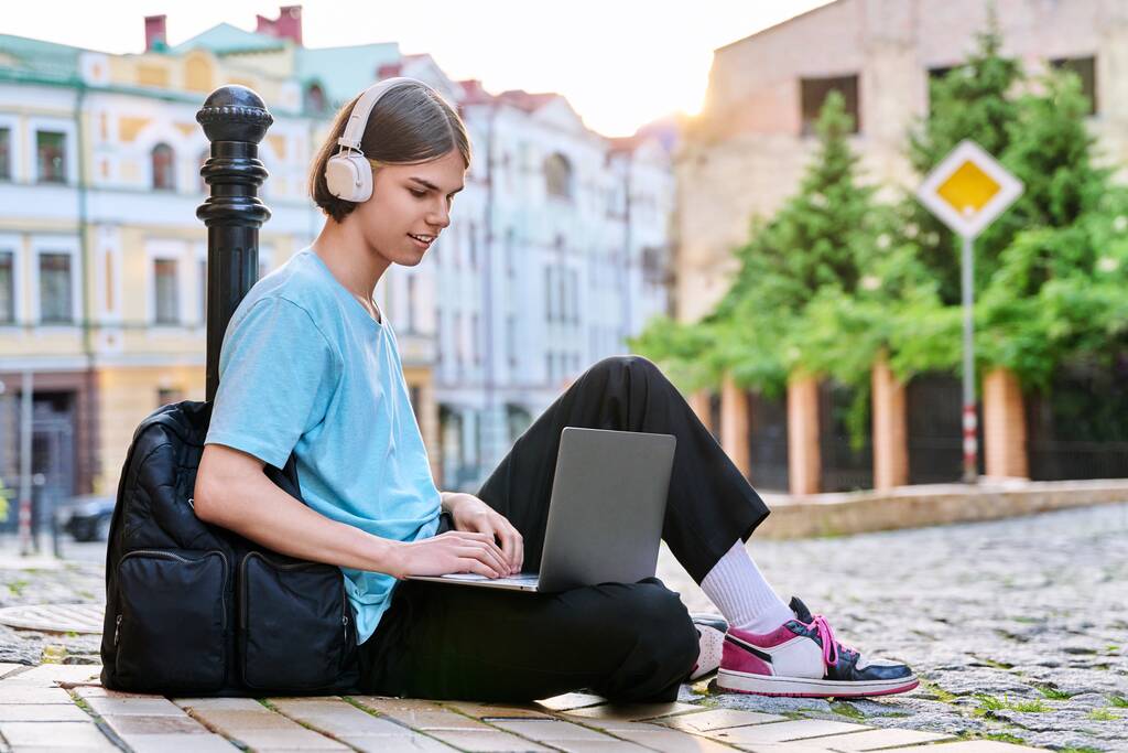 Teenager mit Kopfhörern und Laptop im Freien, auf der Straße der Stadt. Guy 17, 18 Jahre alt, schaut auf den Bildschirm, sitzt auf dem Bürgersteig. College, High School, Jugend, Bildung, Technologiekonzept - Foto, Bild