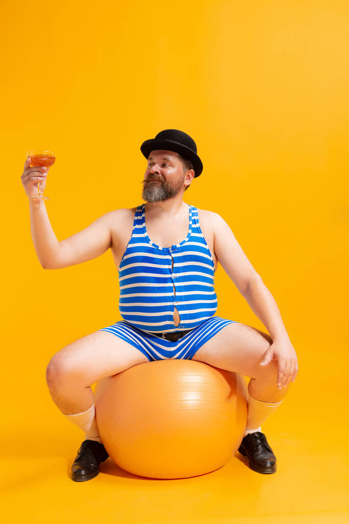 海のビーチで。レトロな縞模様の水着と明るい黄色の背景に隔離された巨大なボールとヴィンテージのボウラー帽子を身に着けているかわいい脂肪の若い男。休暇、夏、面白いミームの感情の概念. - 写真・画像