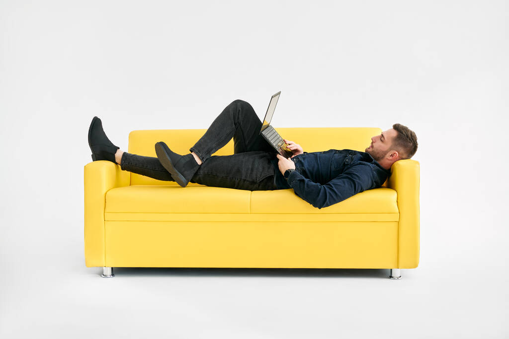 Uomo rilassato sdraiato sul divano giallo utilizzando il computer portatile su sfondo bianco - Foto, immagini