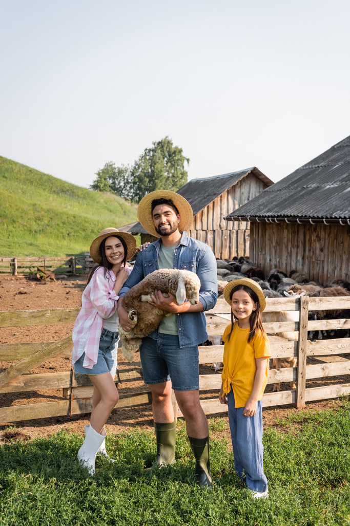 αγρότης με αρνί στο χέρι χαμογελώντας στην κάμερα μαζί με την οικογένεια στο αγρόκτημα στην ύπαιθρο - Φωτογραφία, εικόνα