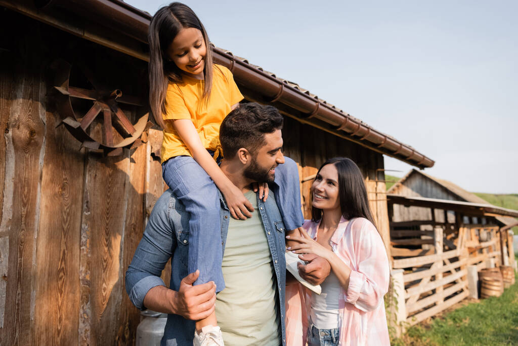 szczęśliwy człowiek piggybacking córka w pobliżu żony i drewniane stodoła na farmie - Zdjęcie, obraz