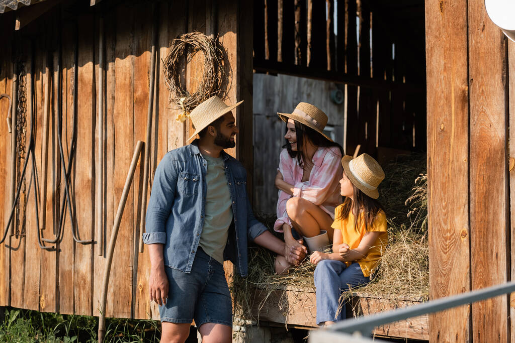 hombre en sombrero de paja hablando con la esposa y la hija sentado en el heno en el granero de madera - Foto, Imagen