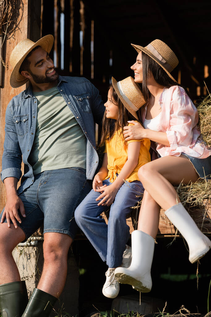 cheerful couple in straw hats looking at each other near daughter on hay in barn - Foto, Imagen