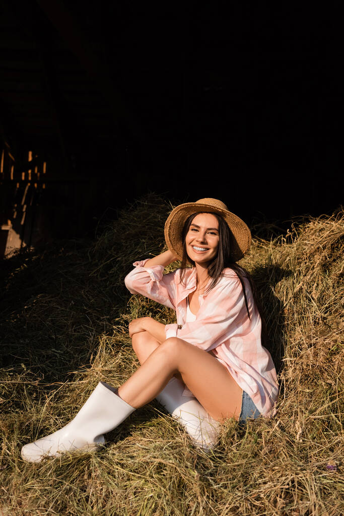 happy farm woman in straw hat sitting on haystack and looking at camera - Zdjęcie, obraz