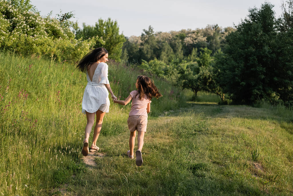 back view of mother and daughter holding hands while running on grassy road near forest - Foto, immagini