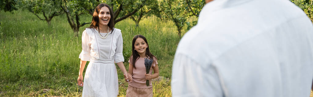 joyful mother and daughter holding hands and smiling near blurred man outdoors, banner - Foto, imagen