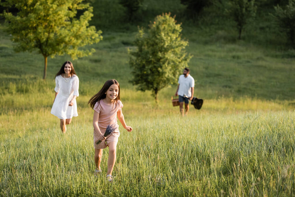 glückliches Mädchen mit Blumenstrauß läuft auf der grünen Wiese in der Nähe von Eltern, die auf verschwommenem Hintergrund gehen - Foto, Bild