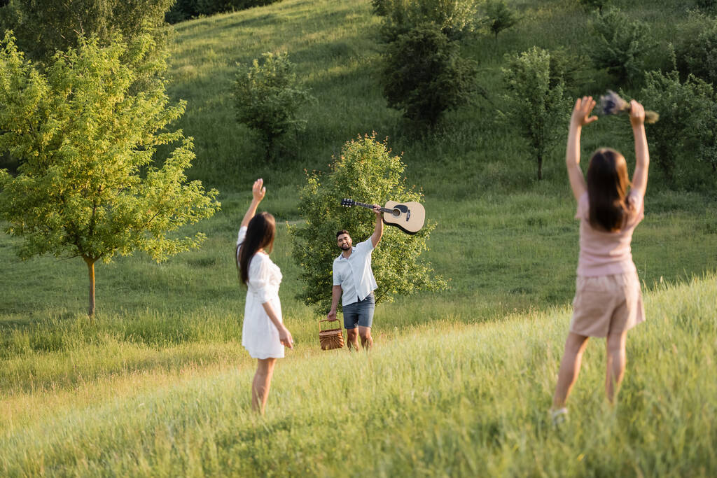 Kadın ve kız çimenli yamaçta akustik gitarlı mutlu adama el sallıyor. - Fotoğraf, Görsel