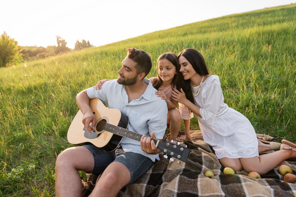 joyful woman and child near man playing guitar on picnic in countryside - Foto, imagen