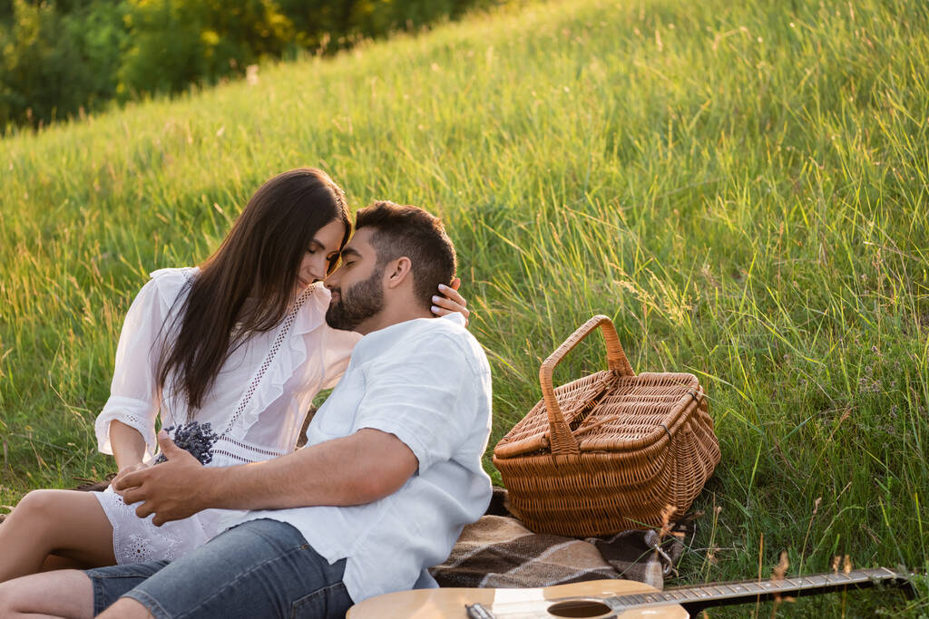 happy couple with closed eyes embracing on lawn near acoustic guitar and wicker basket - Photo, Image