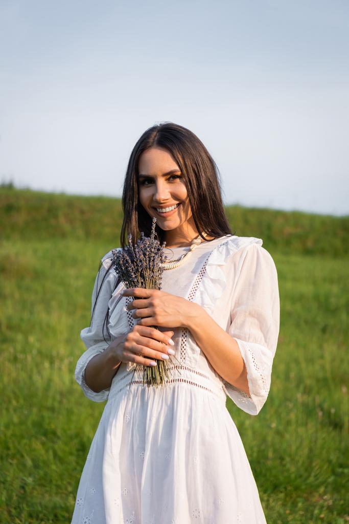 brunette woman in white dress holding lavender bouquet and smiling at camera outdoors - Photo, image