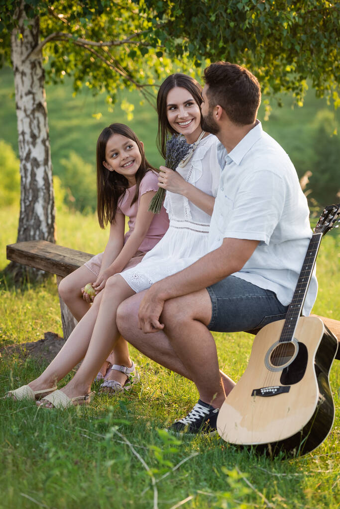 szczęśliwa rodzina rozmawia na ławce obok gitary akustycznej w letni dzień - Zdjęcie, obraz