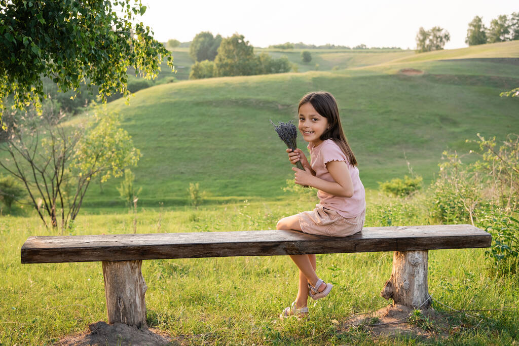 fröhliches Mädchen mit Lavendelblüten, das auf einer Bank auf der Wiese sitzt und in die Kamera blickt - Foto, Bild