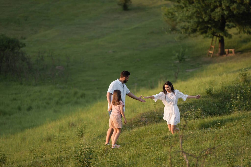 woman extending hand while running on grassy slope towards family - Photo, Image