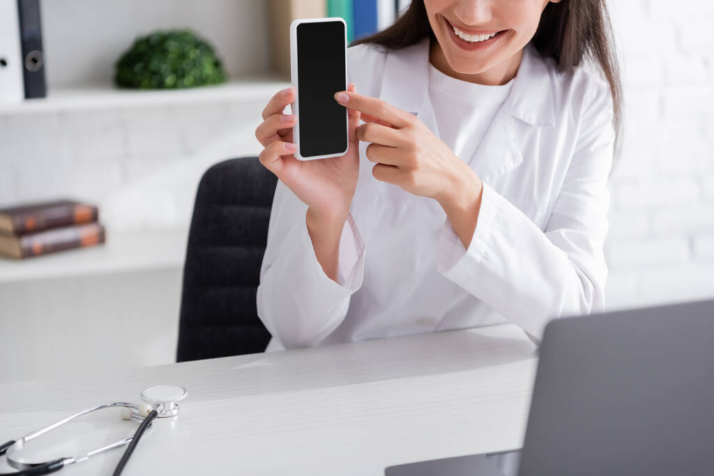 Cropped view of smiling doctor pointing at smartphone during video call on laptop in clinic  - Photo, Image