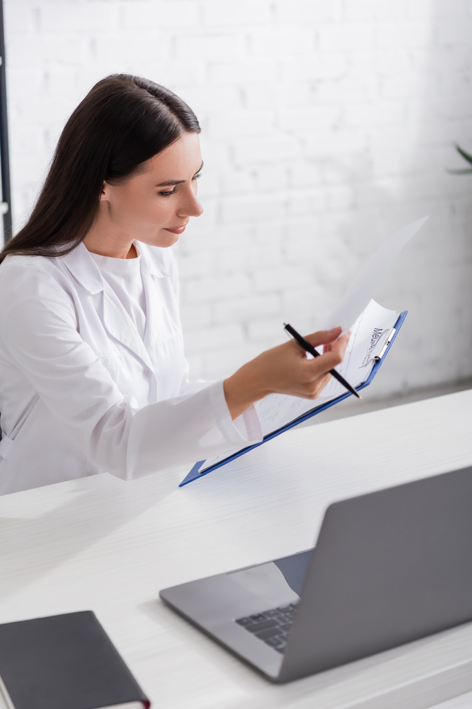 Brunetka lekarz trzymając schowek w pobliżu laptopa i notebooka w klinice  - Zdjęcie, obraz