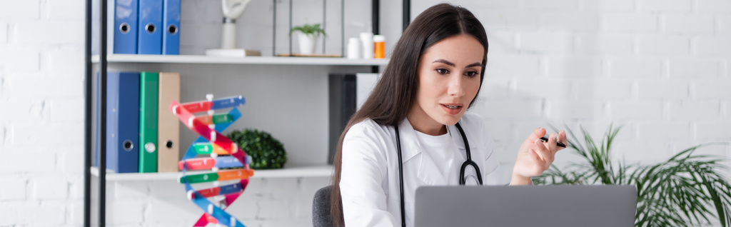 Brunette doctor having video chat on laptop near blurred dna model in clinic, banner  - Photo, Image