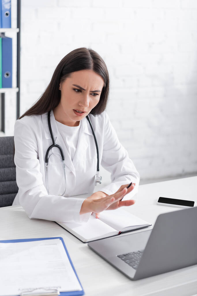 Занепокоєний лікар має відеодзвінок на ноутбук біля буфера обміну інформацією та ноутбука в клініці
  - Фото, зображення