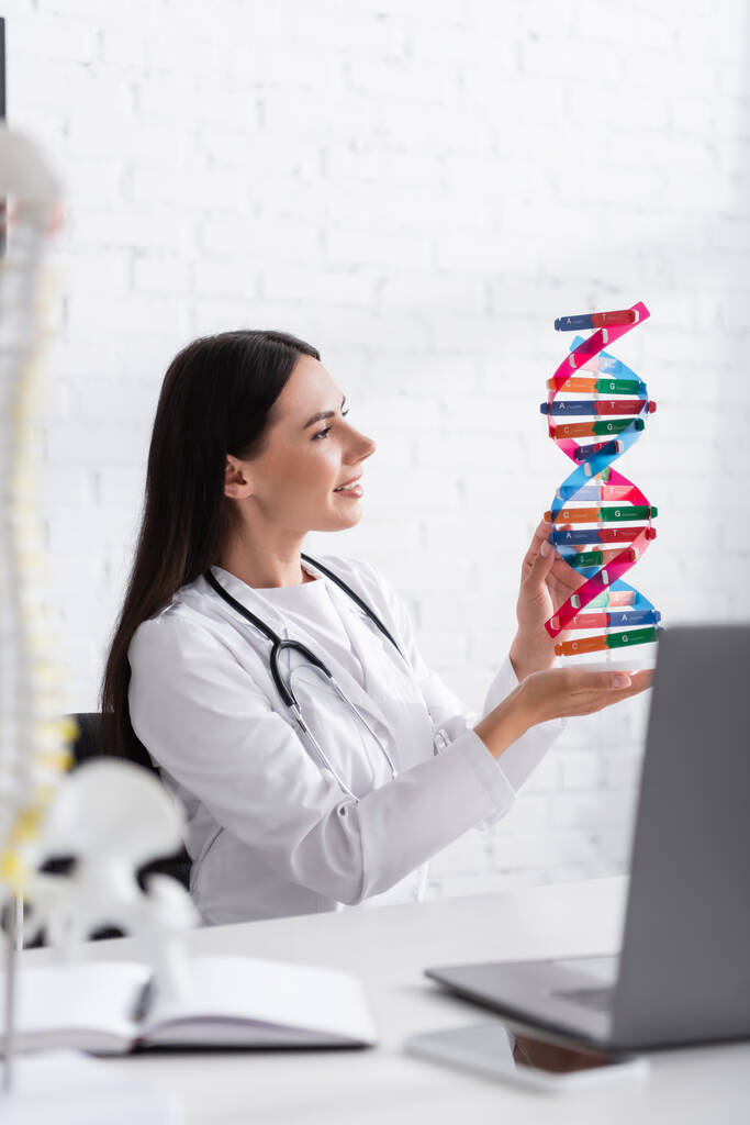 Side view of cheerful doctor holding dna model near blurred gadgets and notebook in clinic  - 写真・画像