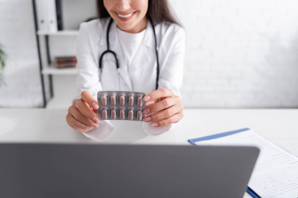 Ausgeschnittene Ansicht eines lächelnden Arztes mit Blister mit Pillen in der Nähe von Laptop und Klemmbrett in der Klinik  - Foto, Bild