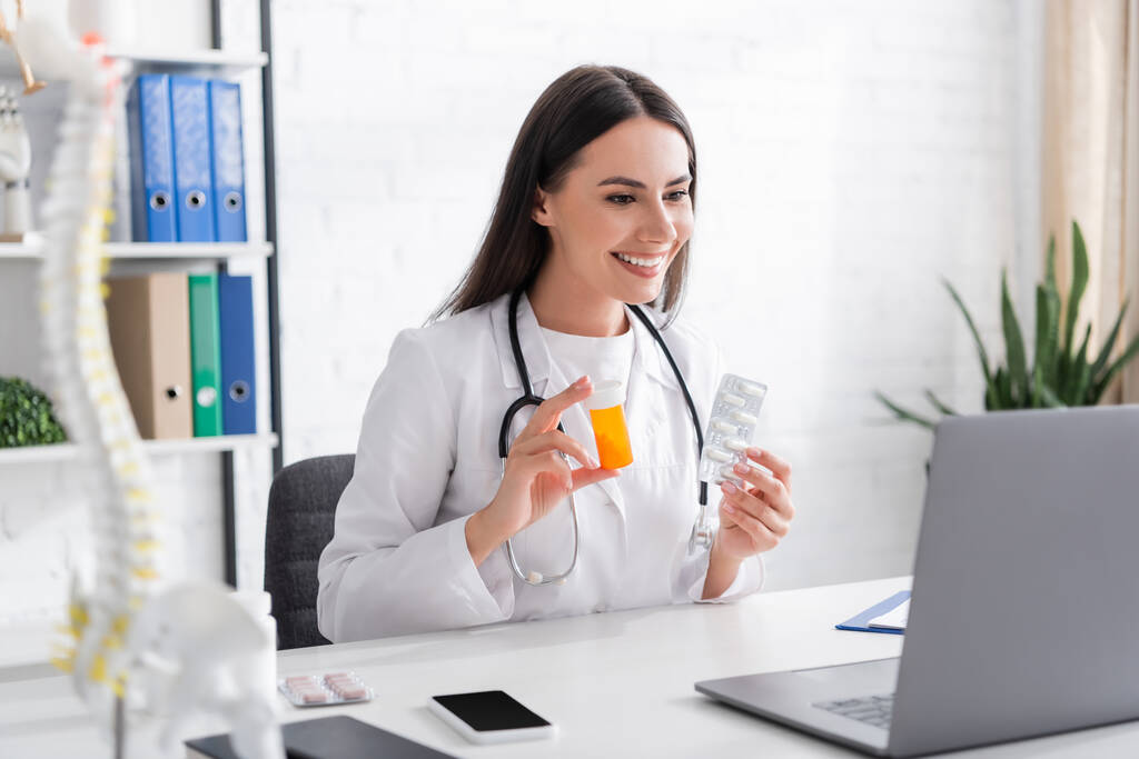 Médico sonriente sosteniendo pastillas y mirando a la computadora portátil durante la videollamada en la clínica - Foto, Imagen