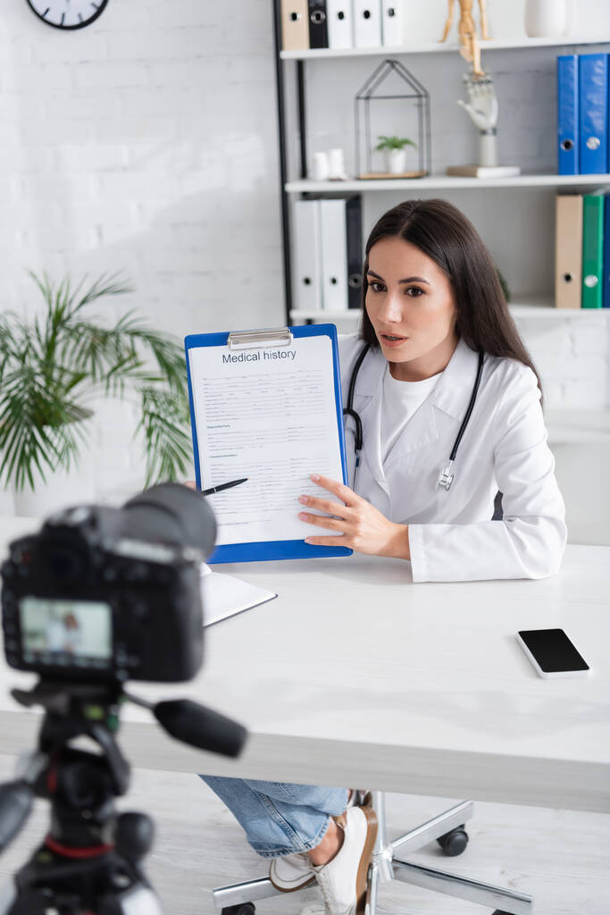 Doctor holding medical history near blurred digital camera in clinic  - Photo, Image