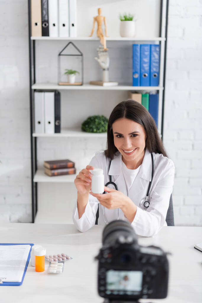 Glimlachende arts met pillen in de buurt van klembord en wazig digitale camera in de kliniek  - Foto, afbeelding