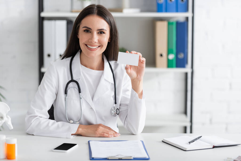 Glimlachende arts met visitekaartje met kopieerruimte bij smartphone en klembord in kliniek  - Foto, afbeelding