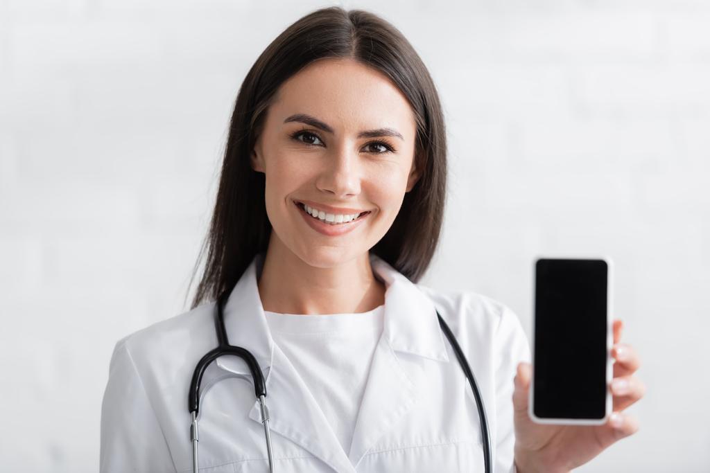 Feliz morena médico sosteniendo smartphone con pantalla en blanco  - Foto, imagen