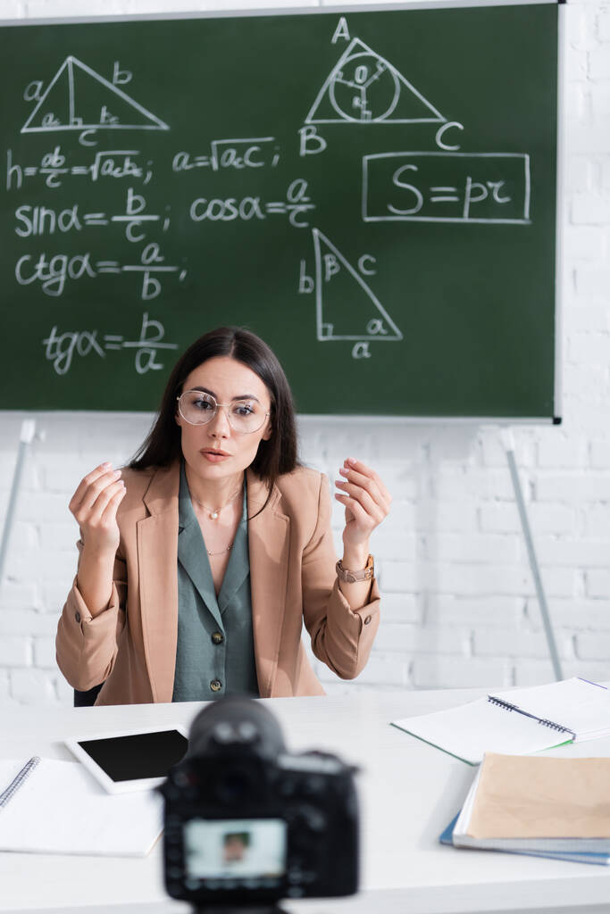 Teacher talking near blurred digital camera and chalkboard with math formulas in classroom  - Photo, image