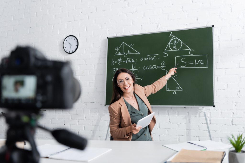 Ευτυχής δάσκαλος κρατώντας ψηφιακή ταμπλέτα και δείχνοντας μαθηματικούς τύπους στο chalkboard κοντά στην κάμερα στο σχολείο  - Φωτογραφία, εικόνα