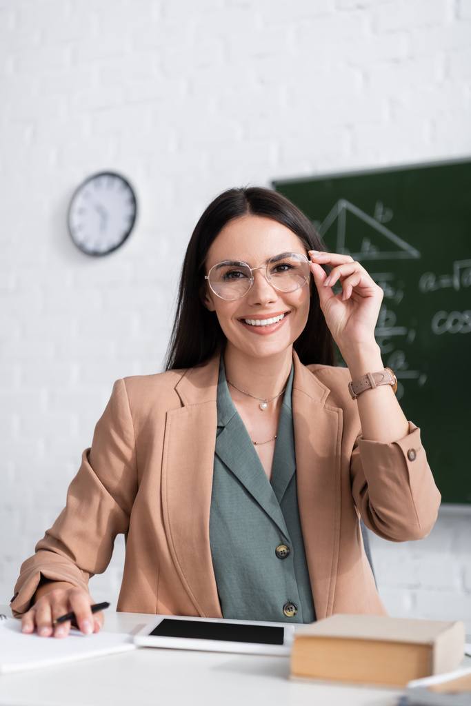 Positive teacher in eyeglasses sitting near digital tablet and book in classroom  - Valokuva, kuva