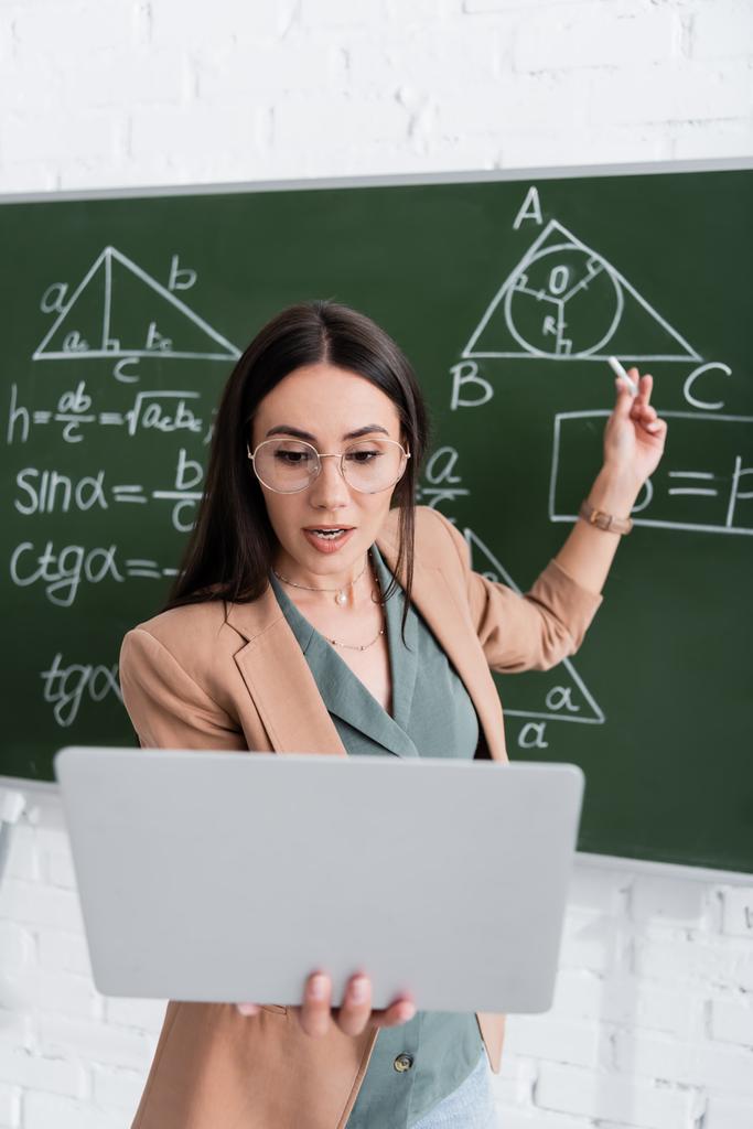 Lehrer mit Videoanruf auf Laptop neben Tafel mit Mathe-Formeln  - Foto, Bild