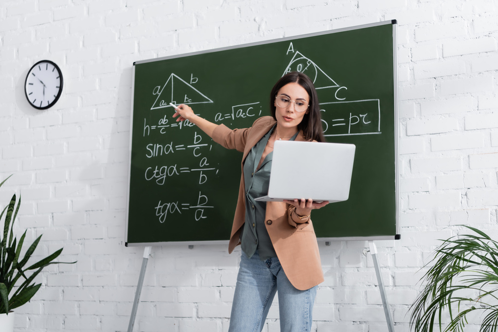 Учитель указывает на математические формулы на доске и держит ноутбук во время онлайн урока в классе  - Фото, изображение