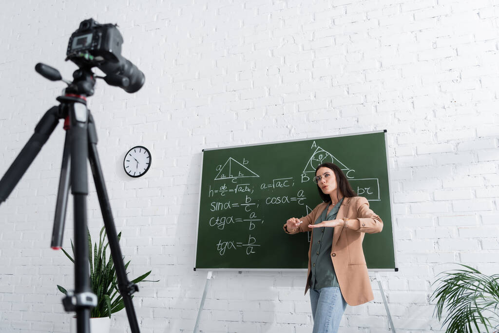 Nauczyciel gestykulujący w pobliżu tablicy z formułami matematycznymi i aparatem cyfrowym w klasie  - Zdjęcie, obraz