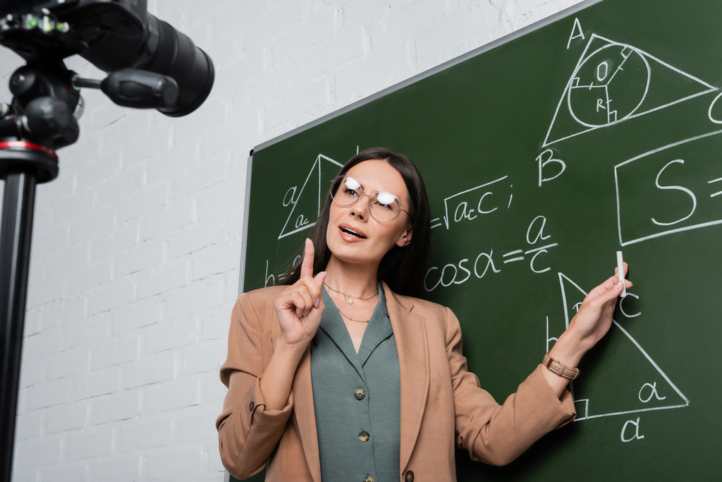 Tanár szemüveges magyarázza matematikai képletek táblán közelében digitális fényképezőgép osztály  - Fotó, kép