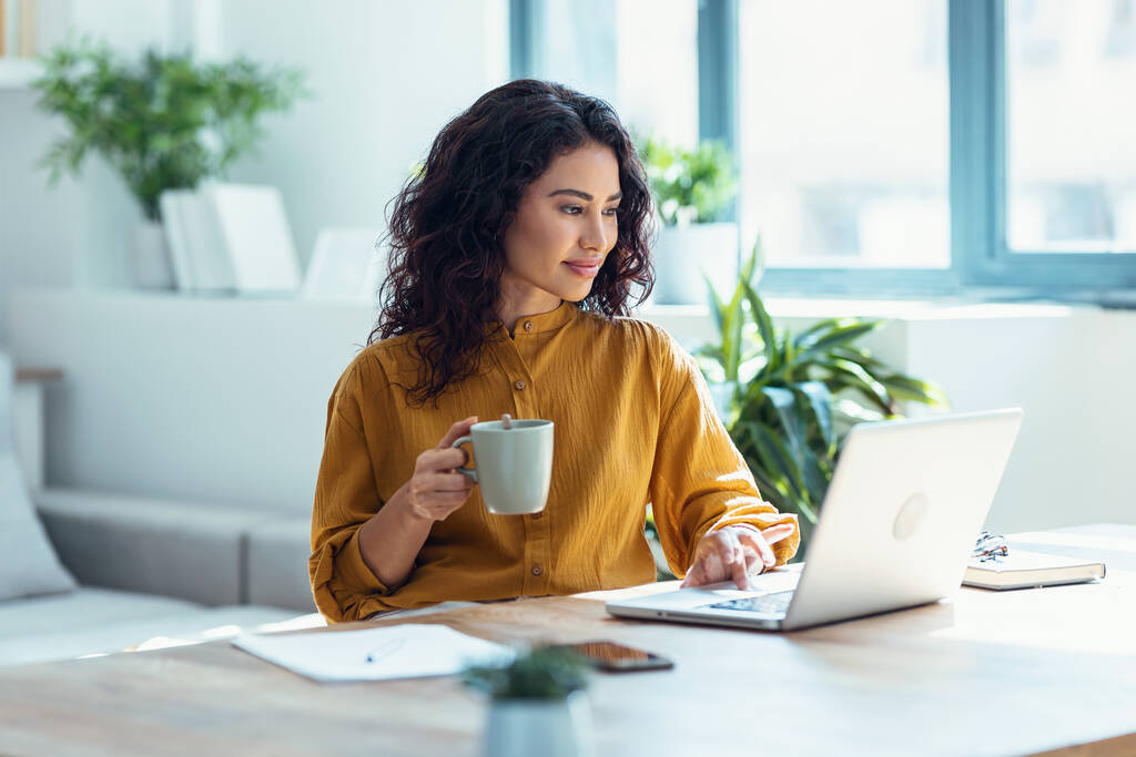 Снимок сконцентрированной красивой деловой женщины, работающей с ноутбуком, когда она пьет кофе в гостиной дома. - Фото, изображение