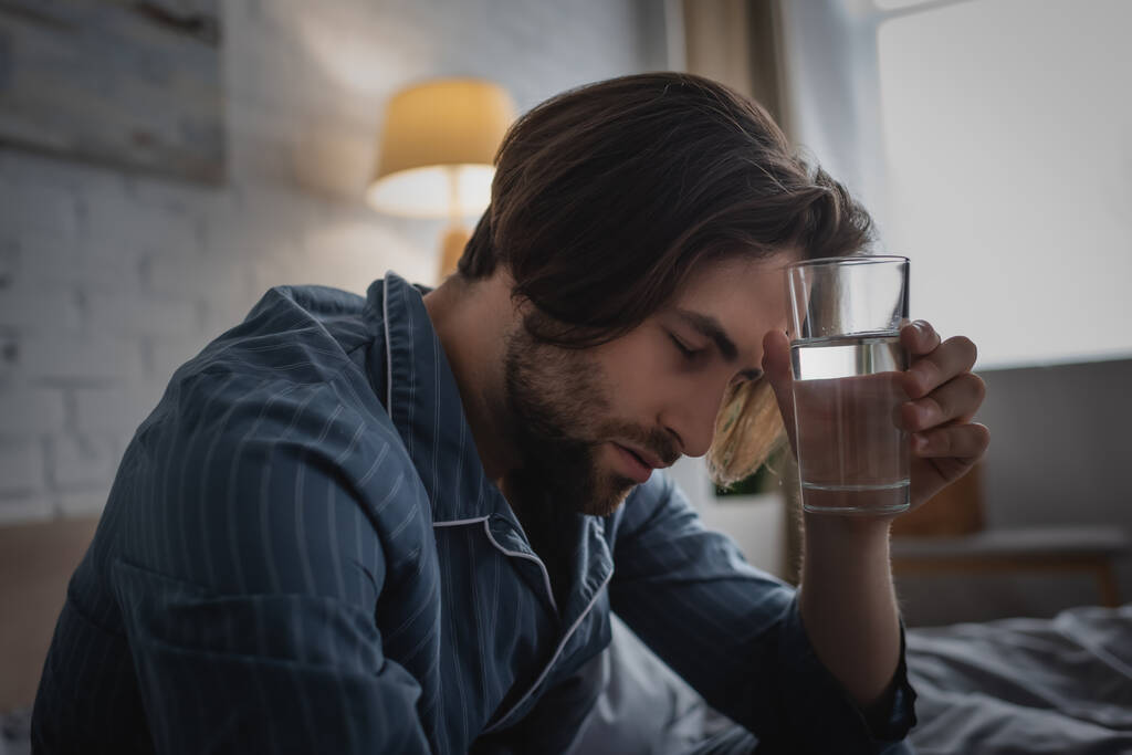 Tired man in pajama holding glass of water in bed in morning  - Zdjęcie, obraz