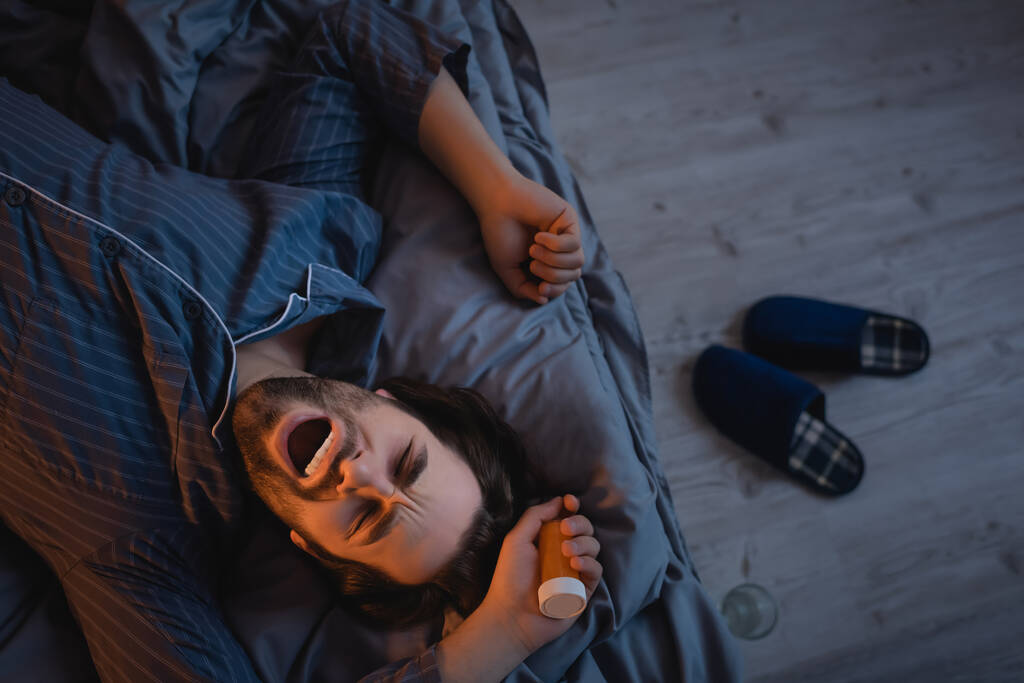 Top view of man with insomnia holding pills and yawning on bed  - Valokuva, kuva