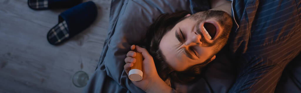 Top view of bearded man yawning and holding pills on bed at night, banner  - Foto, afbeelding