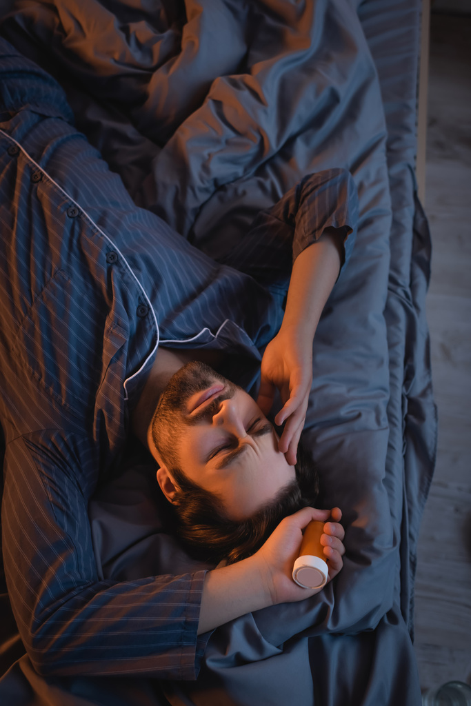 Вид зверху на втомленого чоловіка з безсонням тримає таблетки і торкається голови на ліжку
  - Фото, зображення