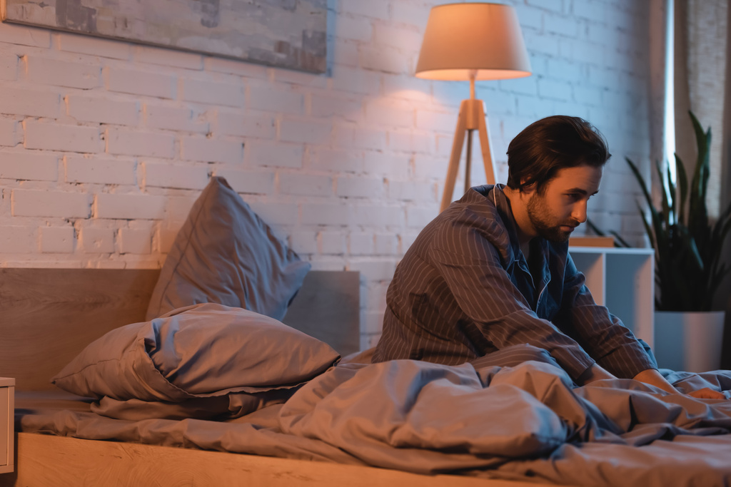 man met baard zittend op bed terwijl hij 's nachts aan slaapstoornis lijdt  - Foto, afbeelding