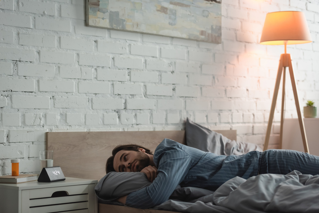 Mladý muž při pohledu na hodiny, zatímco trpí nespavostí v posteli v dopoledních hodinách  - Fotografie, Obrázek
