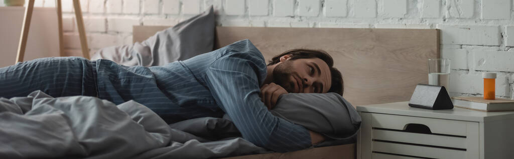 Бородатый мужчина смотрит на будильник возле воды и таблетки на тумбочке в спальне, баннер  - Фото, изображение