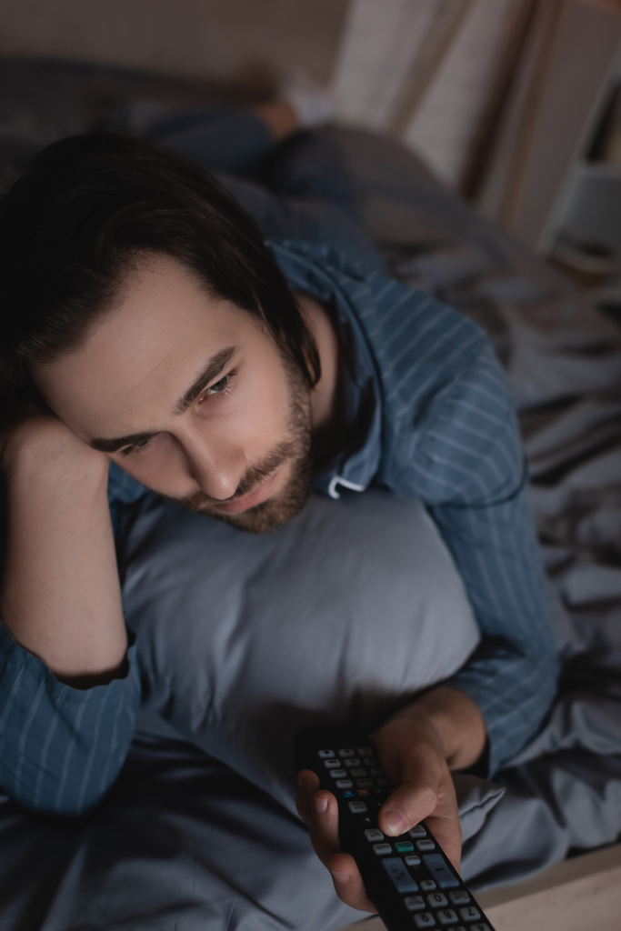 Bearded man clicking channels while lying on bed at night   - Foto, immagini