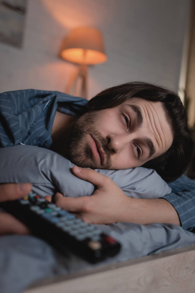 Man with insomnia holding blurred remote controller on bed  - Valokuva, kuva