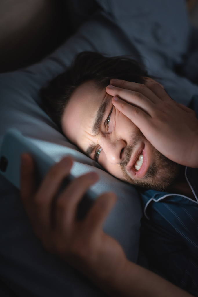 Напряженный человек с бессонницей с помощью размытого смартфона на кровати ночью  - Фото, изображение