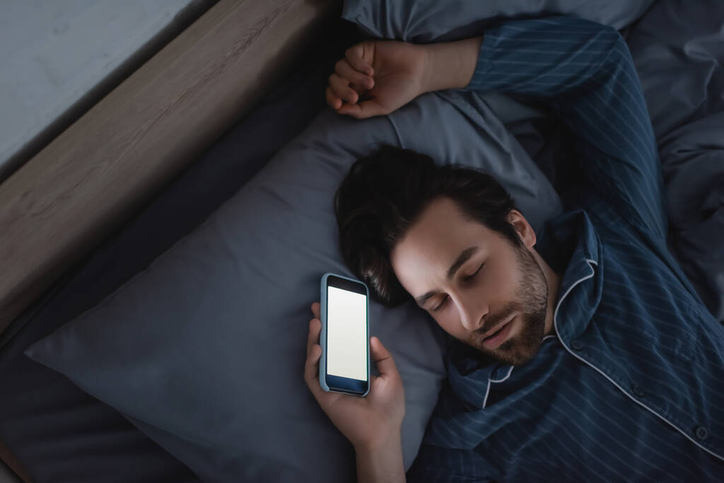 Genç adamın evde uyurken elinde boş ekranla akıllı telefonu tutmasının en üst görüntüsü  - Fotoğraf, Görsel