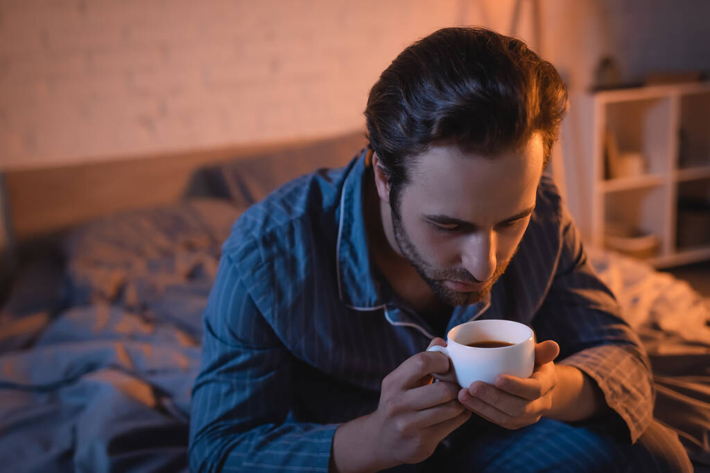 Young man in pajama holding cup of coffee on bed at night  - Fotografie, Obrázek
